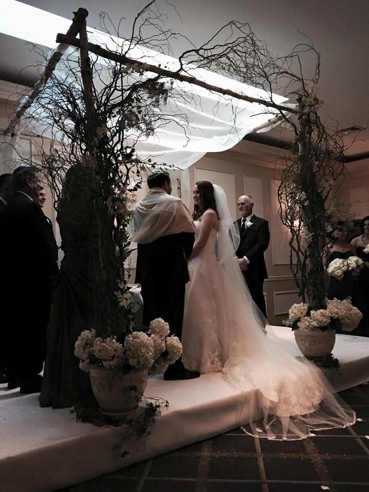 Wedding ceremony under a white canopy with bride, groom, and officiant beside floral arrangements at dusk