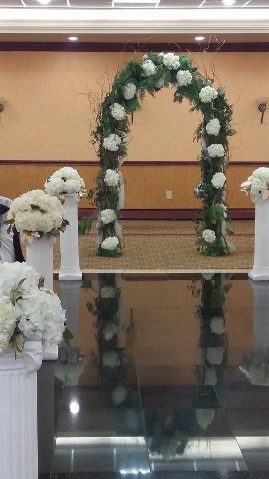 White floral wedding arch with greenery in a banquet hall, flanked by white flower arrangements on pedestals.