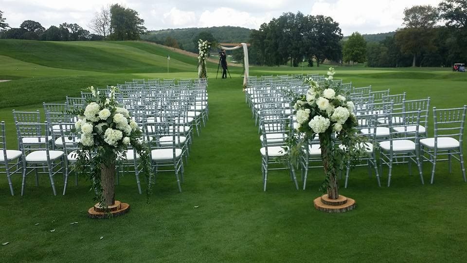Outdoor wedding ceremony setup on a green lawn with white chairs, floral arrangements, and an archway.