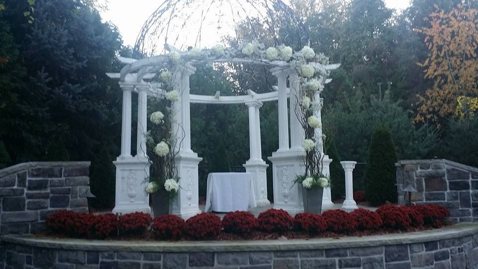 White wedding arch and draped canopy on a stone stage with red flowers in a garden setting