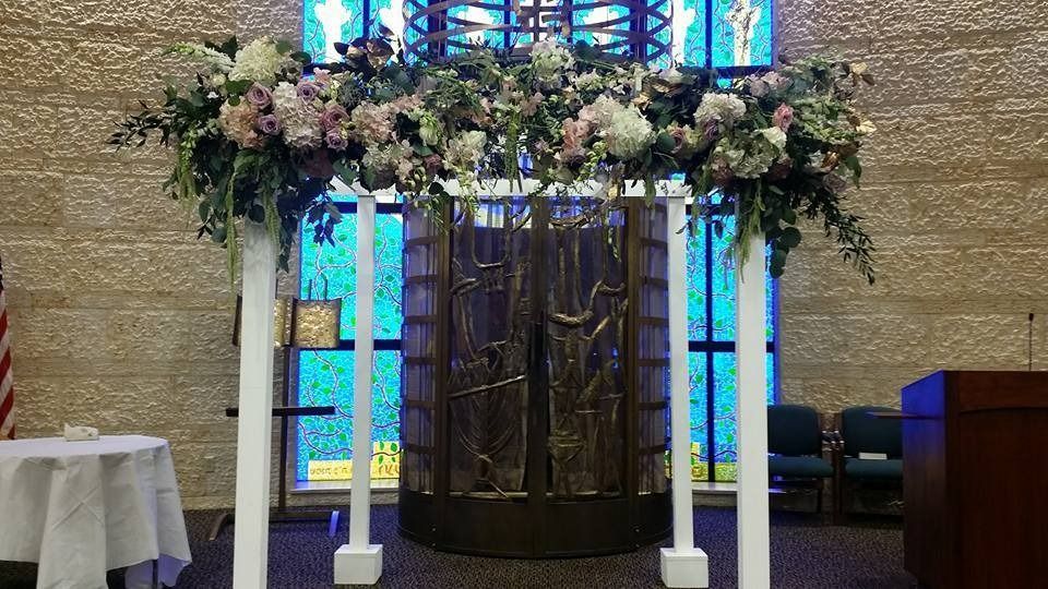 Floral-decorated wedding arch before a wooden menorah, with stained glass windows behind it.