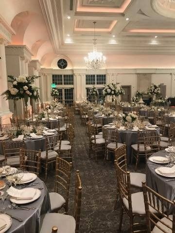 Elegant banquet hall set with round tables, gold chairs, floral centerpieces, and chandeliers under a lit ceiling.