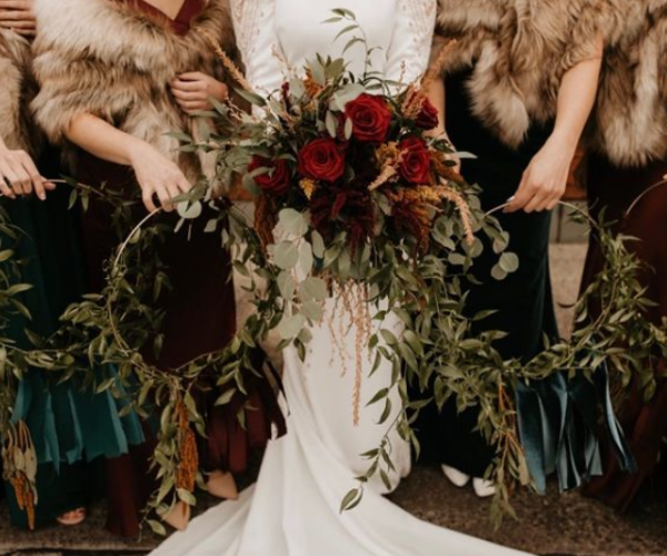 Bridesmaids holding a floral wreath around a white wedding gown with red and green flowers
