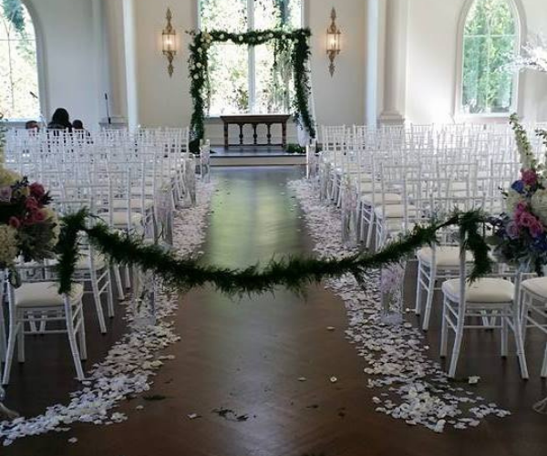 Decorated wedding aisle with white chairs, floral arch, and scattered petals in a bright ceremony hall