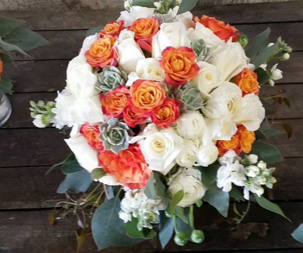 Round bouquet of white, cream, and orange roses with green foliage on a rustic wooden surface