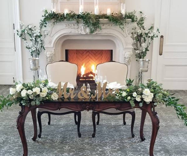 Elegant fireplace with floral garlands, two white chairs, and a decorated table in a bright room