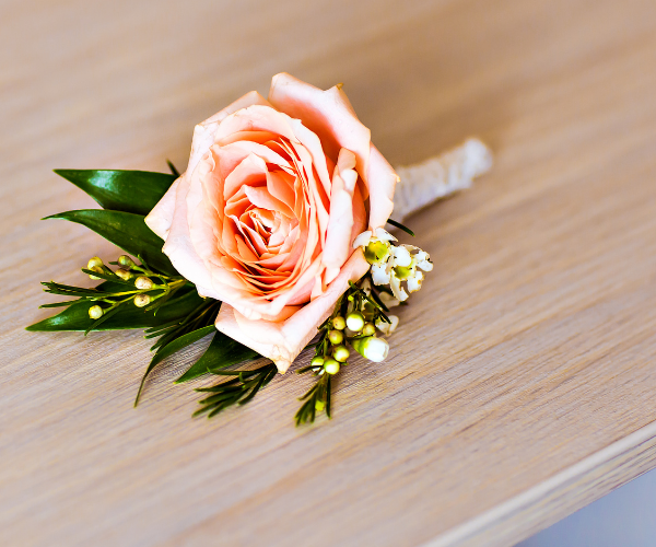 Peach rose boutonniere with green leaves and white filler flowers on a wooden surface