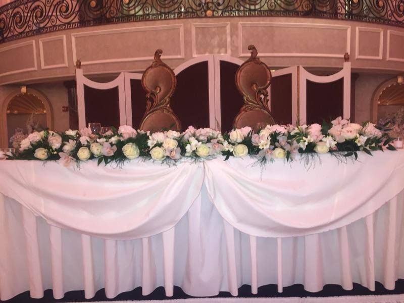 Decorated banquet table with white drapery and pink-and-white floral arrangement, set beneath ornate wooden chairs