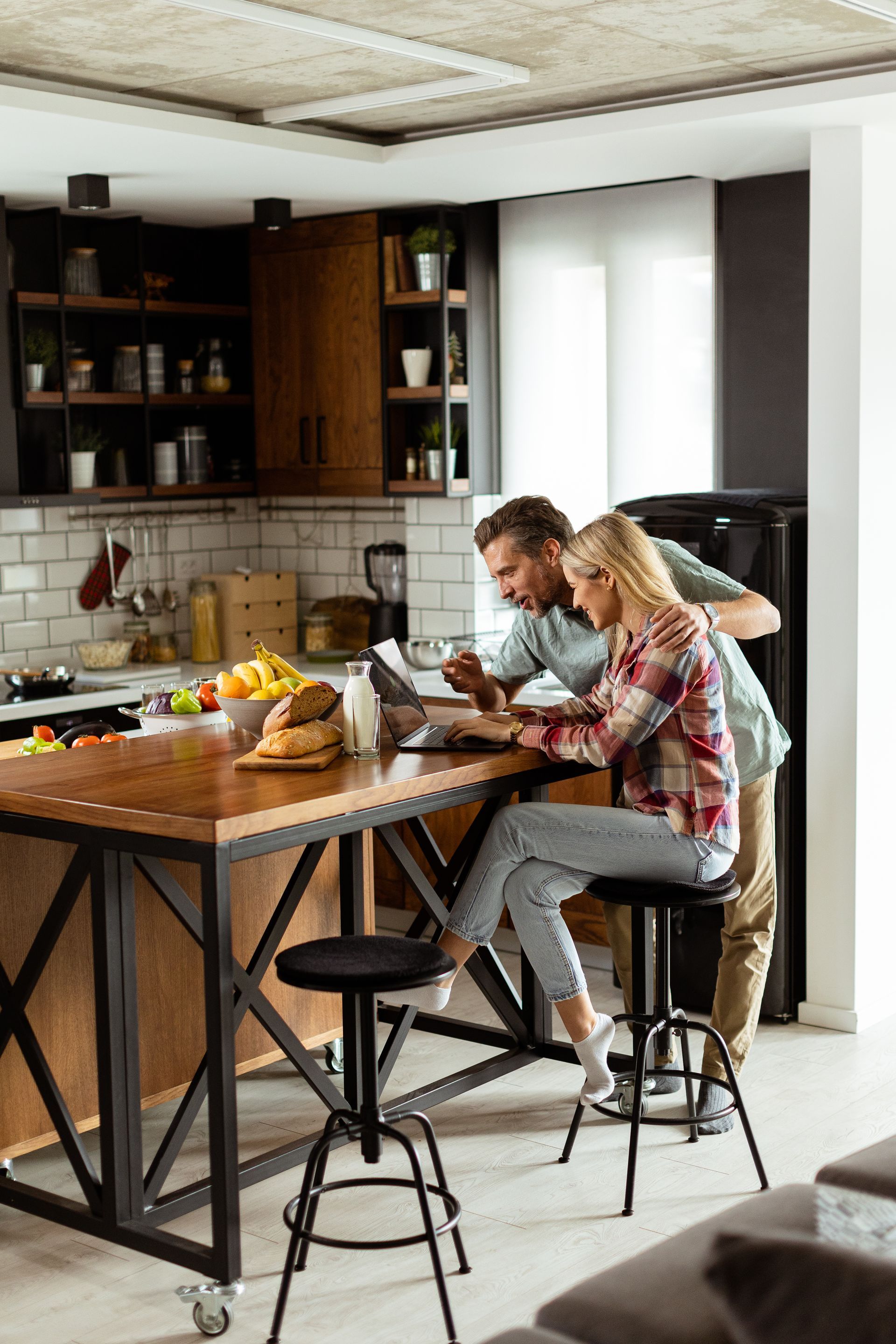 A man and a woman are sitting at a table in a kitchen looking at a laptop.