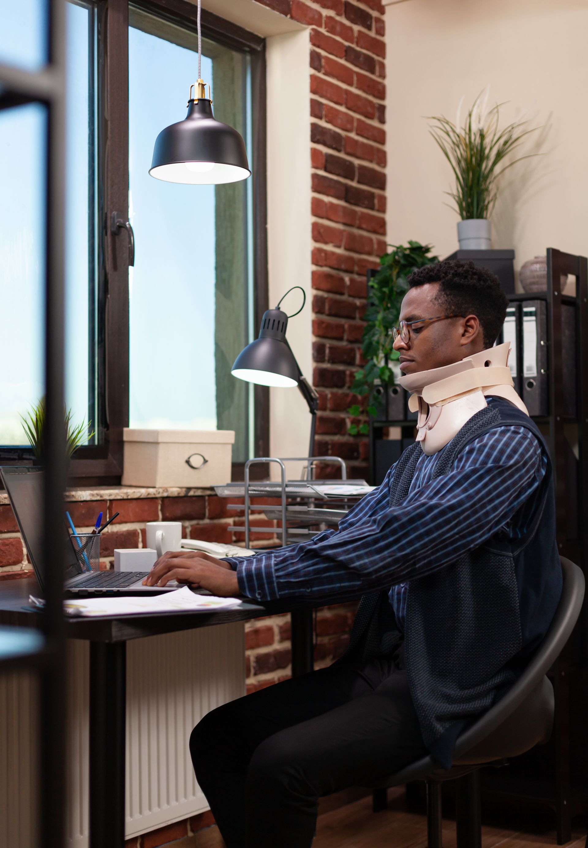 A man with a neck brace is sitting at a desk using a laptop computer.