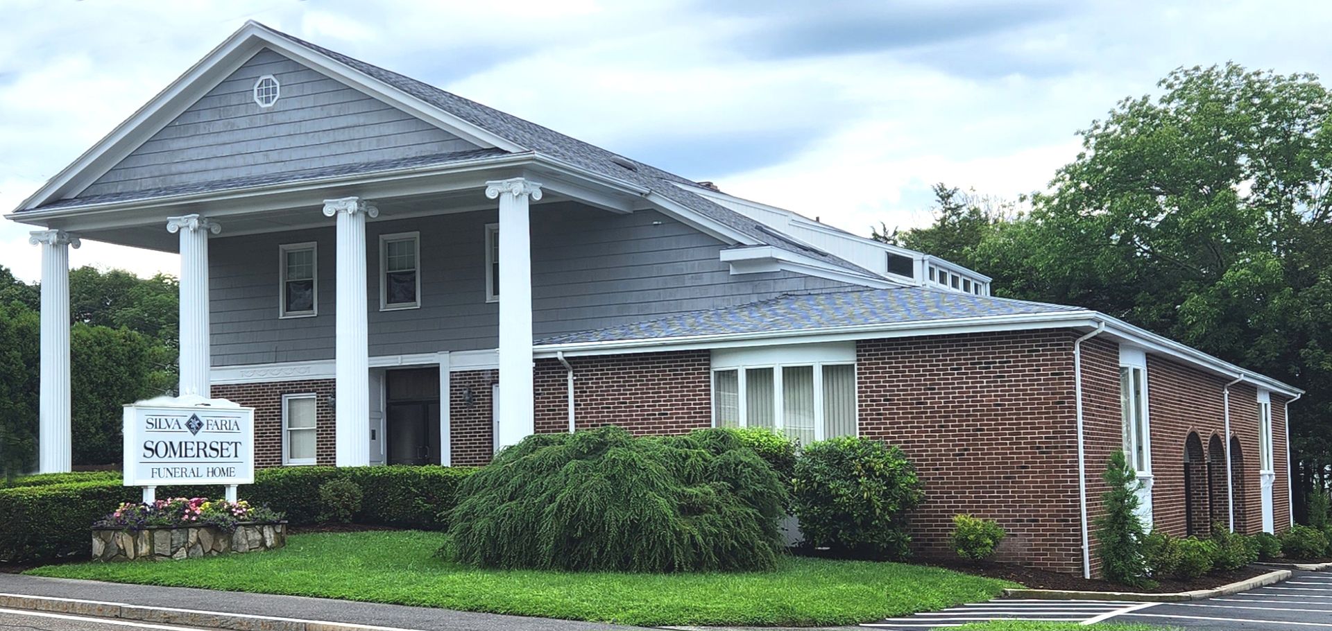 Building with columns and brick siding; sign in front. Green grass and overcast sky.