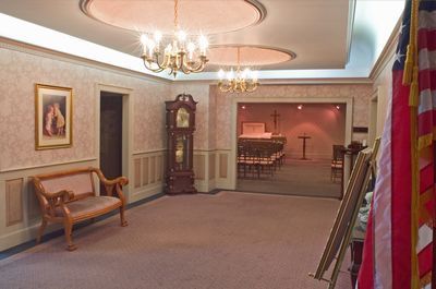 Interior of a funeral home with chandelier lighting, a longcase clock, and a bench; an American flag is on the right.
