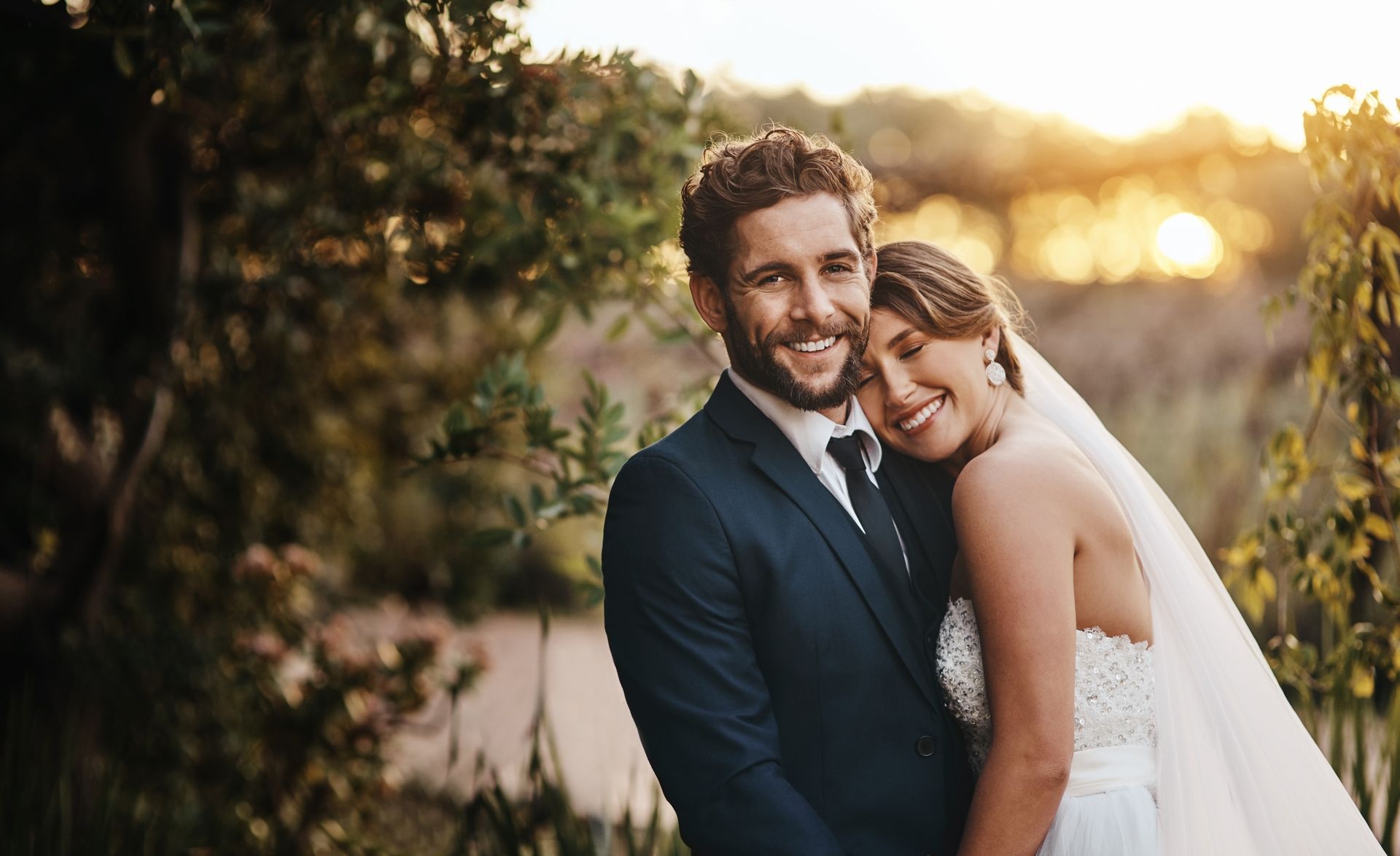 A couple smiles warmly while embracing outdoors at sunset, the bride resting her head on the groom’s shoulder.