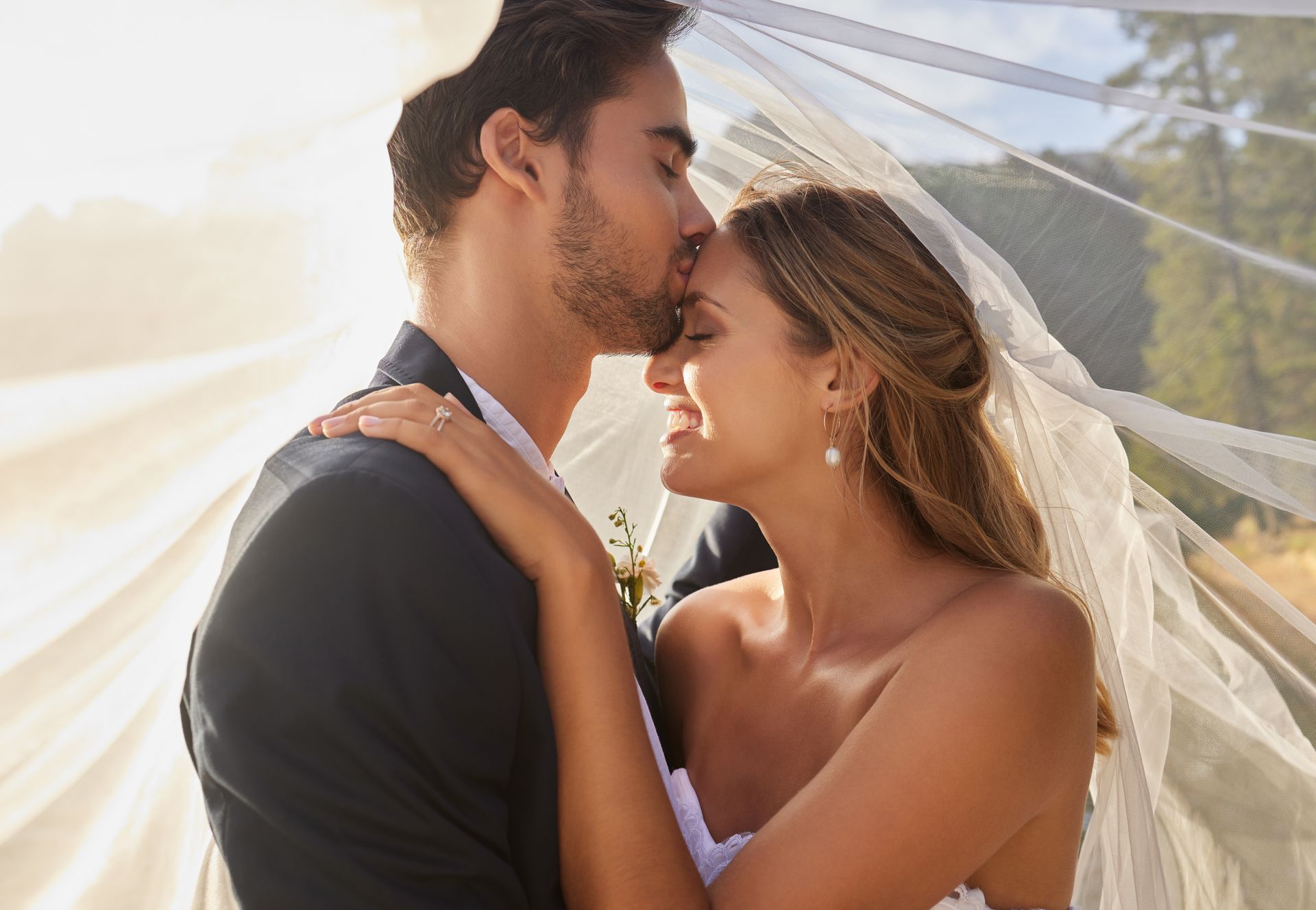 A groom kisses the bride's forehead under a flowing white veil, both smiling softly in a sunlit outdoor setting.