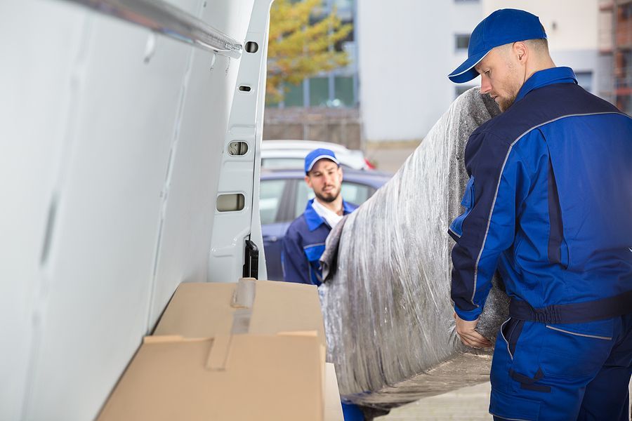 Two professional movers in blue uniforms loading a large, plastic-wrapped sofa into a white delivery van.
