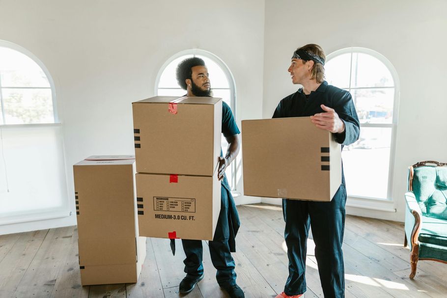 Two movers carrying cardboard boxes in a bright room.