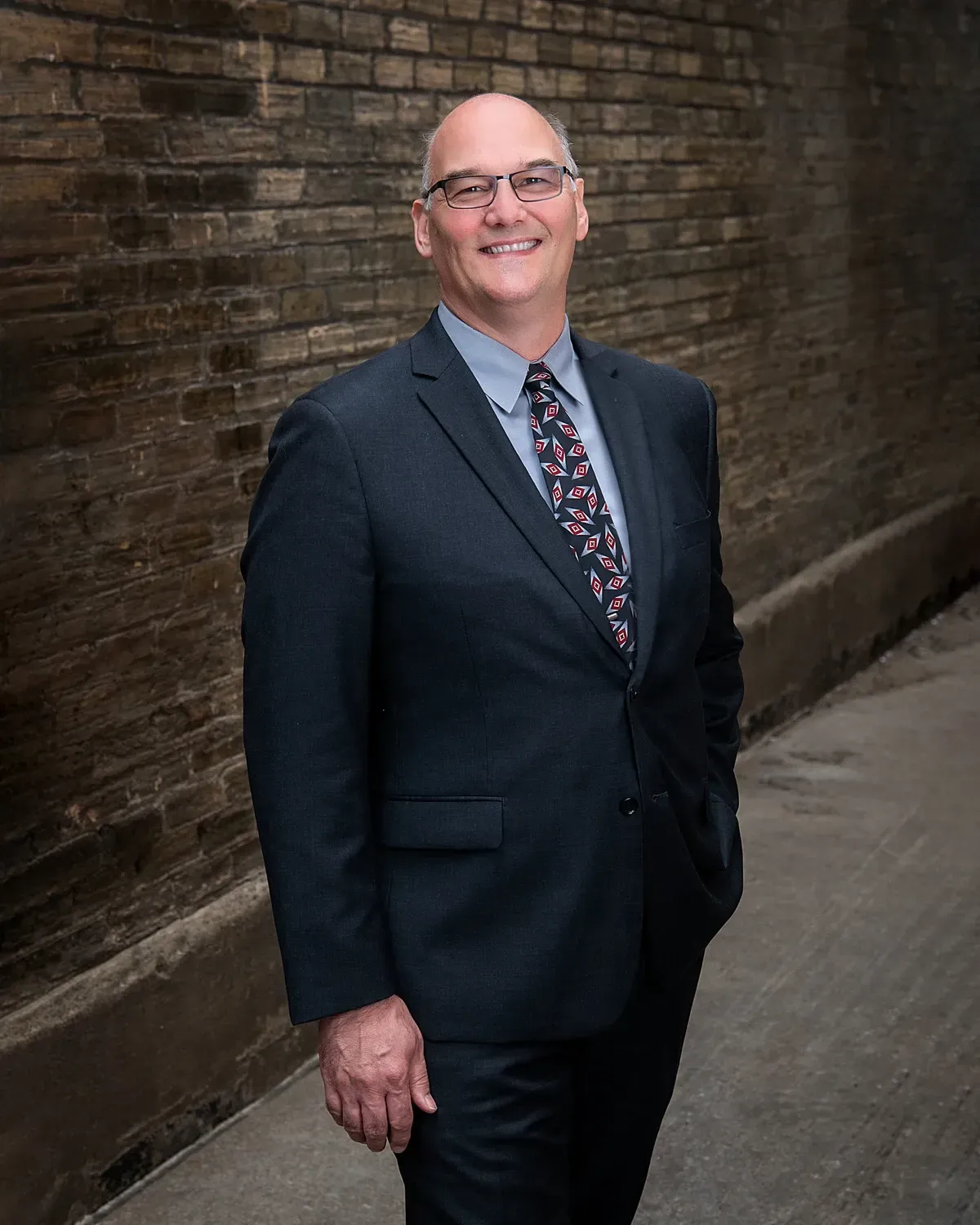 Man in a dark suit smiling beside a brick wall in a narrow alleyway