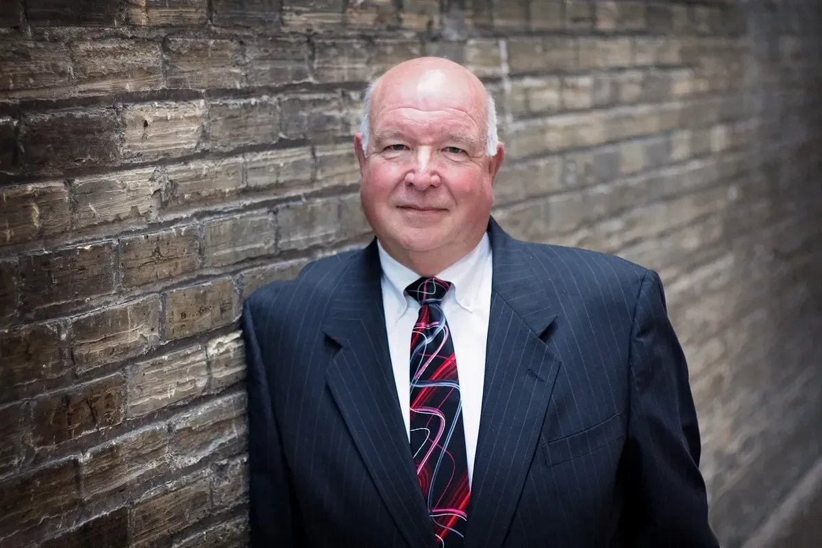 Man in a dark suit and red tie stands against a brick wall, facing the camera.