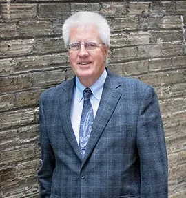 Man in a blue plaid suit jacket standing against a stone wall, smiling at the camera