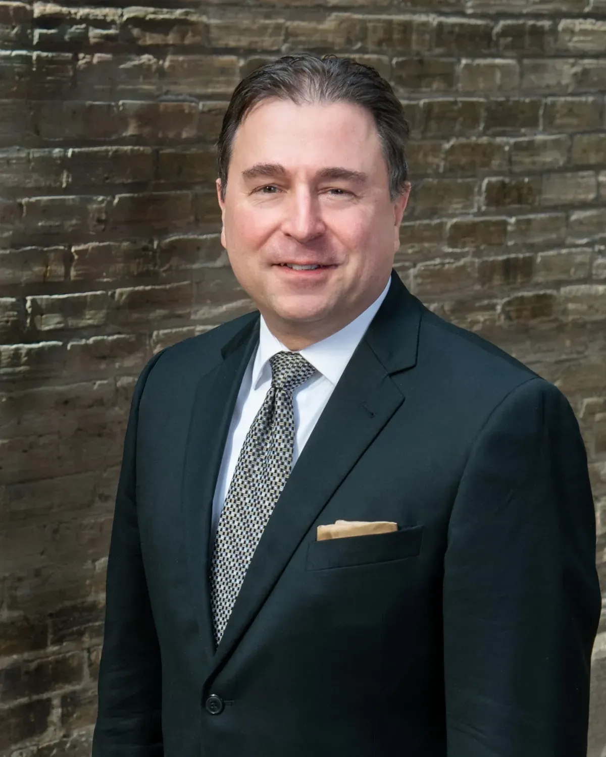 Man in dark suit and patterned tie posing against a brick wall