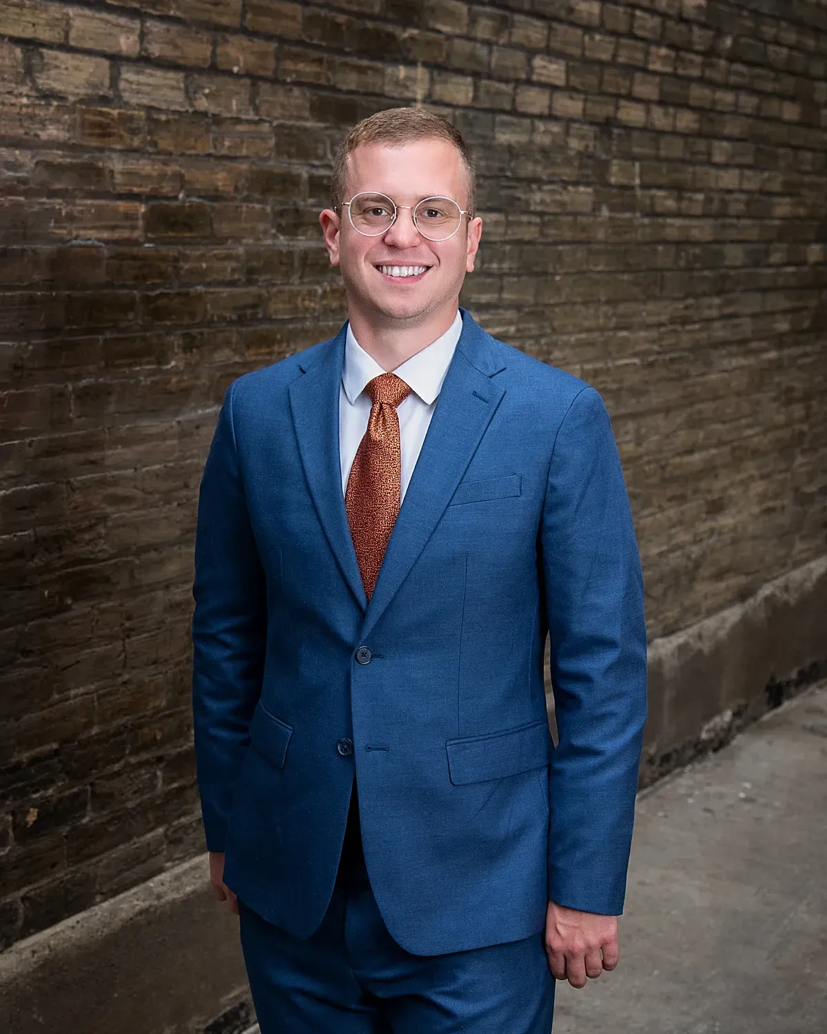 Man in a blue suit and red tie smiling beside a brick wall in an alleyway