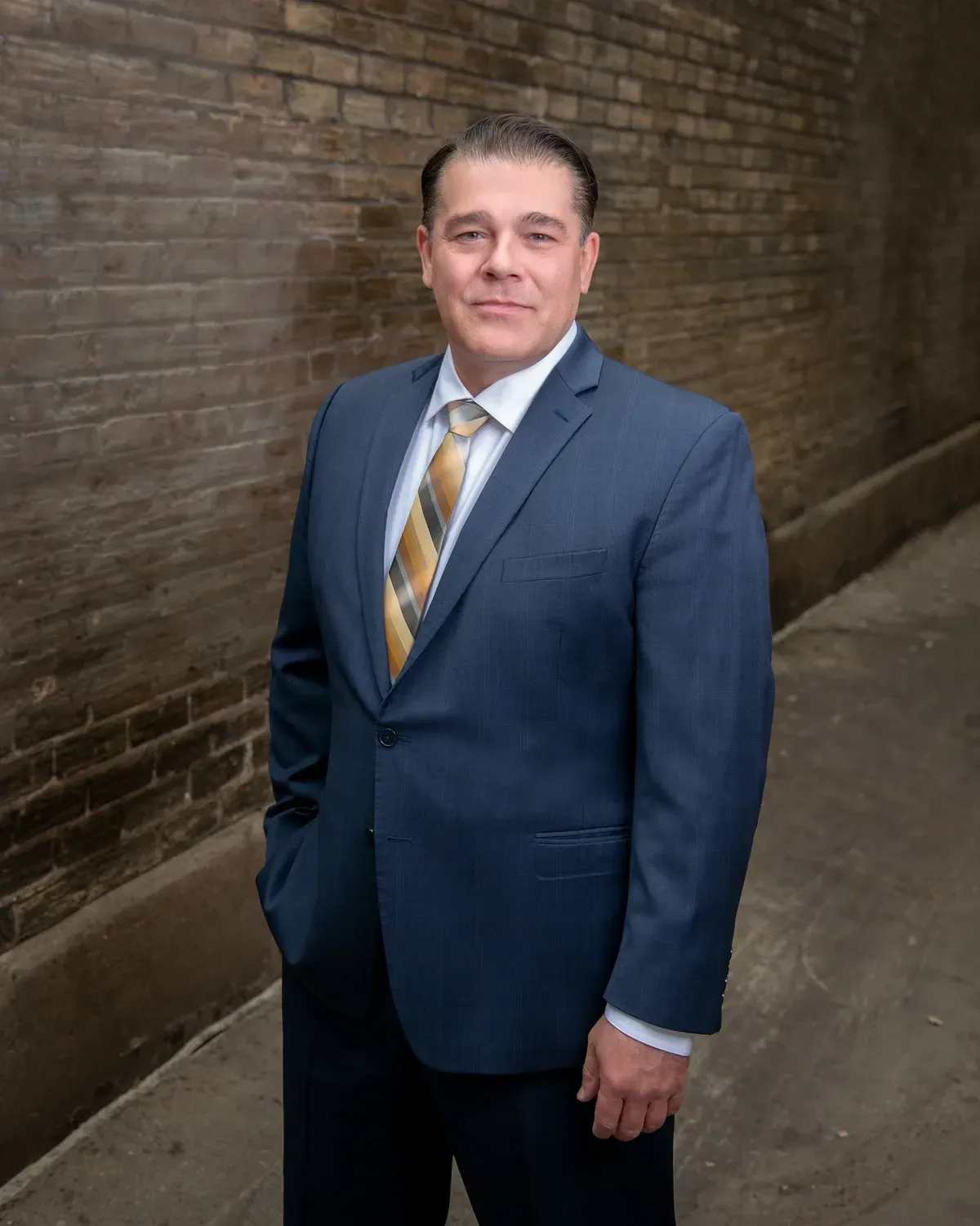 Man in a blue suit and striped tie standing in a narrow brick alleyway.