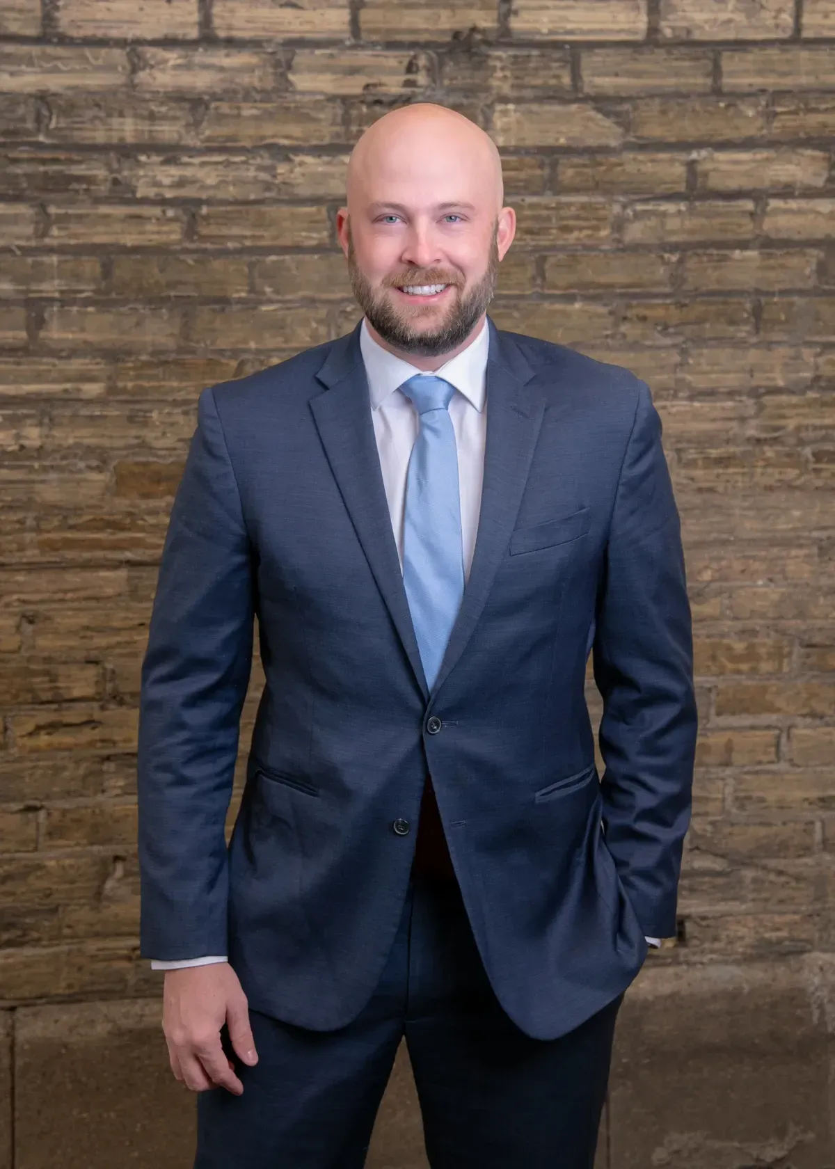 Man in a navy suit and light blue tie stands smiling in front of a brick wall.