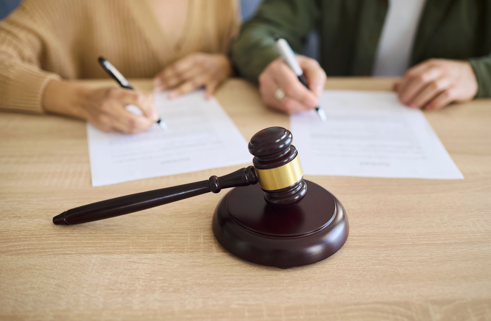 Judge’s gavel on a desk as two people sign legal documents in the background. Judge’s gavel on a desk as two people sign legal documents in the background.