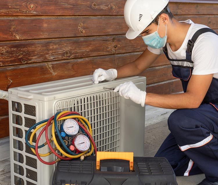 Technician in hard hat and mask inspecting an outdoor air conditioner unit with gauges attached