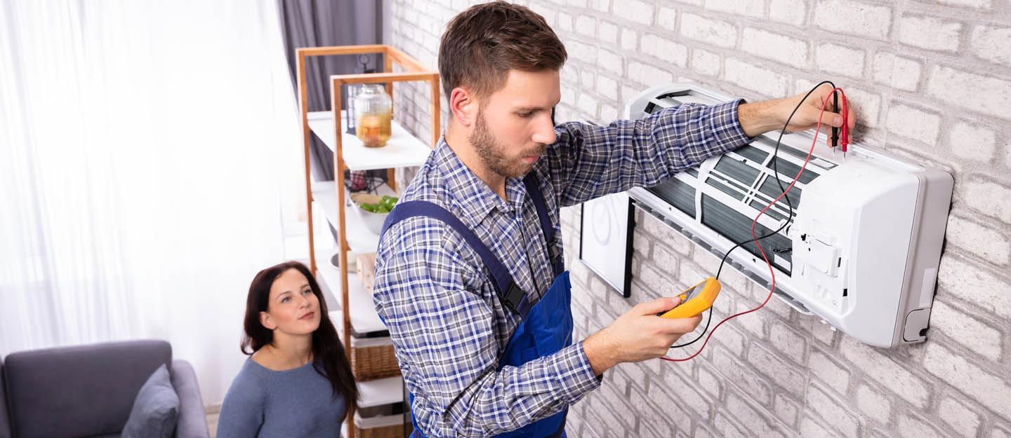Technician repairing a wall-mounted unit while a woman watches from the couch