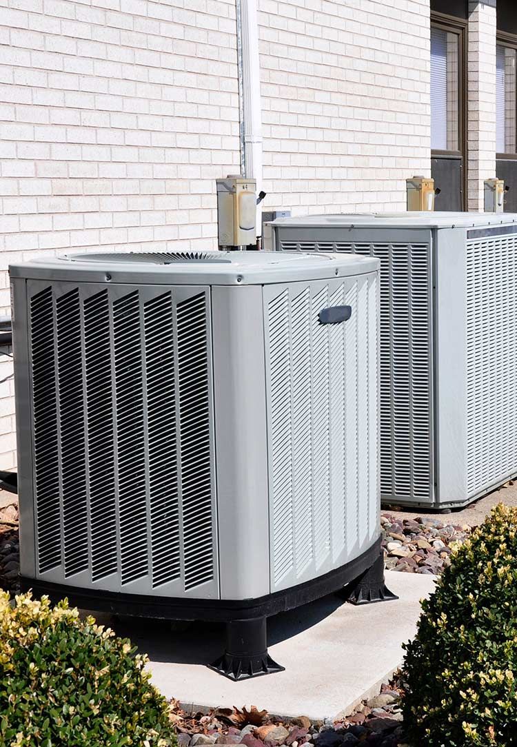 Two large outdoor HVAC units beside a brick building and shrubs