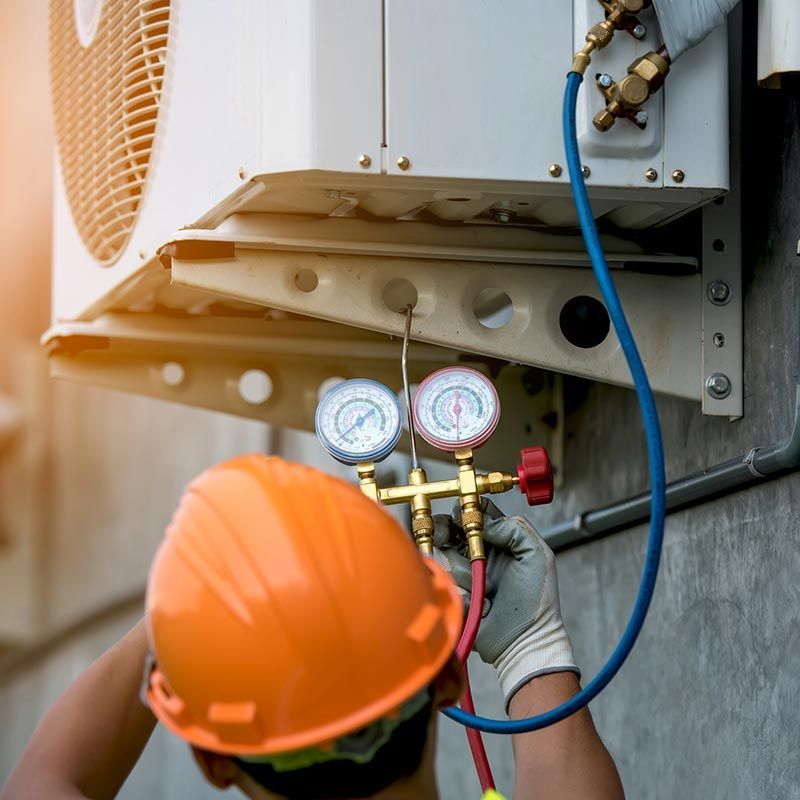 Technician in orange hard hat checking AC unit gauges with blue hose outdoors