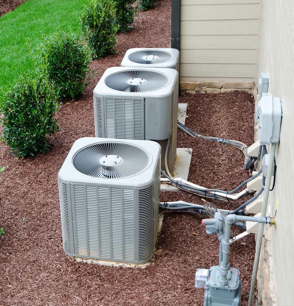 Three outdoor air conditioning units beside a house with hoses and utility lines.