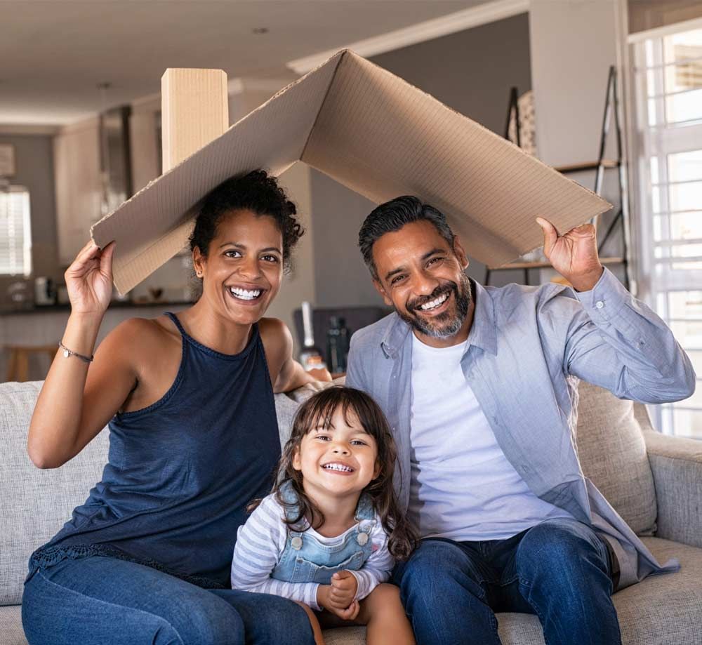 Family smiling under a cardboard roof, sitting together on a couch in a bright living room.