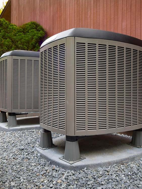 Two large outdoor HVAC units on gravel beside a wooden fence.
