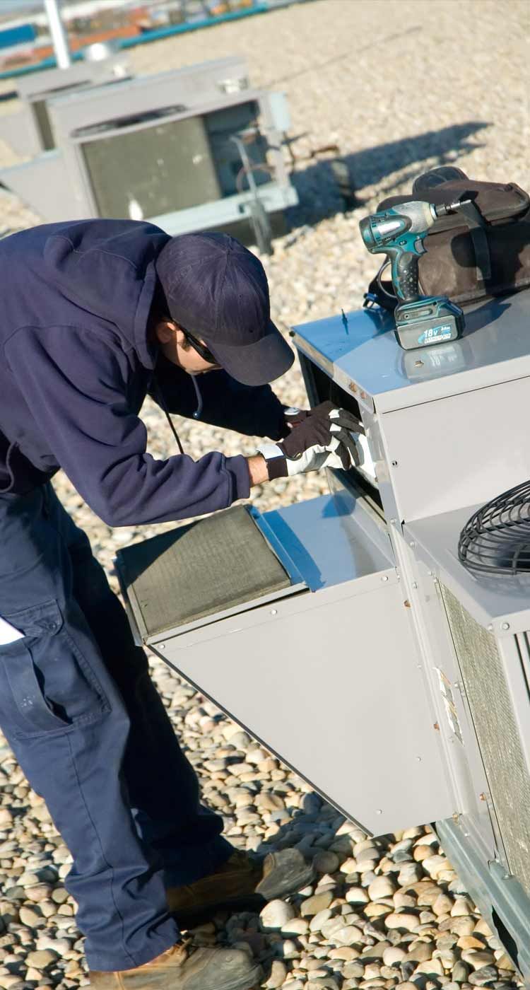Technician in navy workwear repairs rooftop HVAC equipment beside a gray unit.