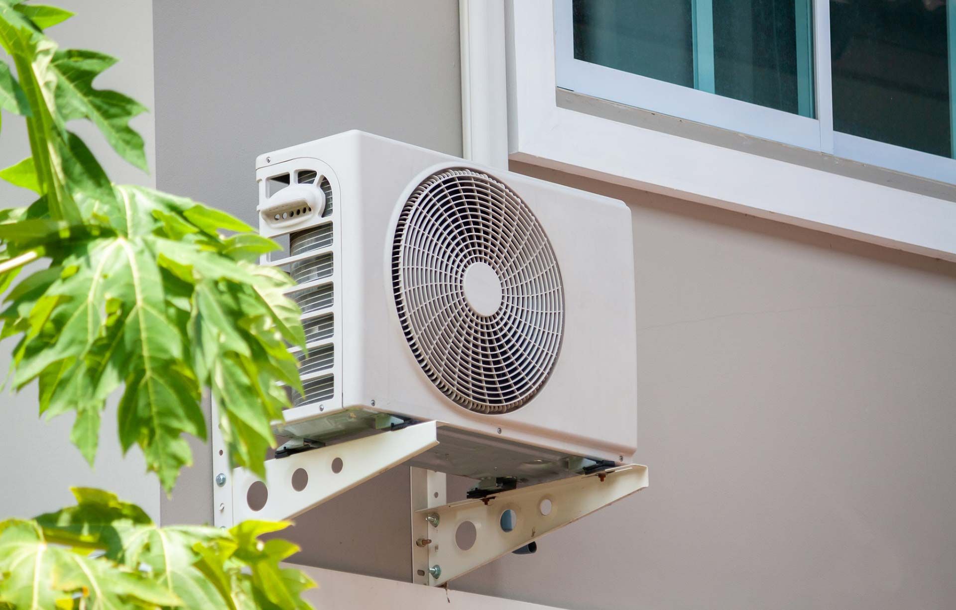 Outdoor wall-mounted air conditioner unit beside a window, partly framed by green leaves.
