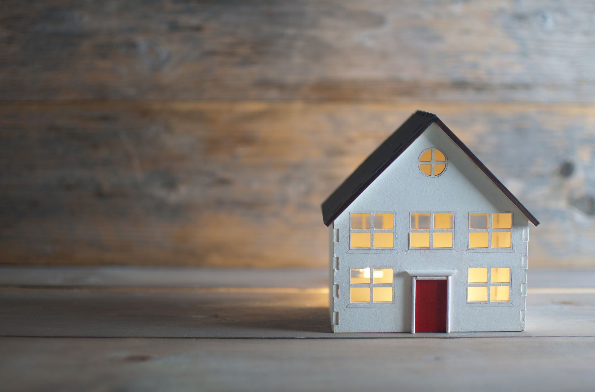 Miniature light-up house with a black roof, white walls, and a red door, set against a wooden background.