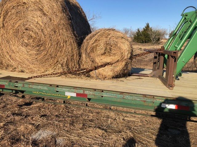 Hay bales on a trailer, one secured with a chain, being loaded with a green tractor.