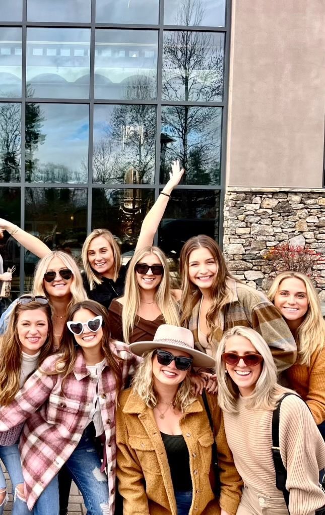 Group of smiling women posing outdoors near a building with large windows.