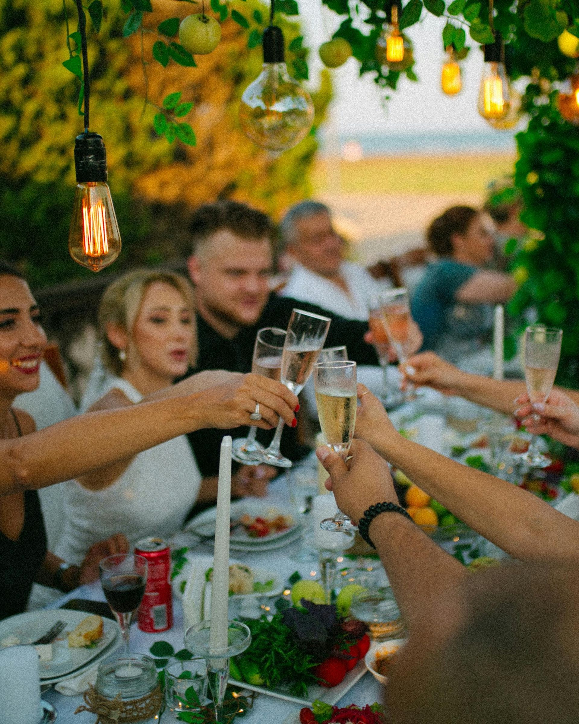 Friends toasting with champagne at a candlelit outdoor dinner table under greenery
