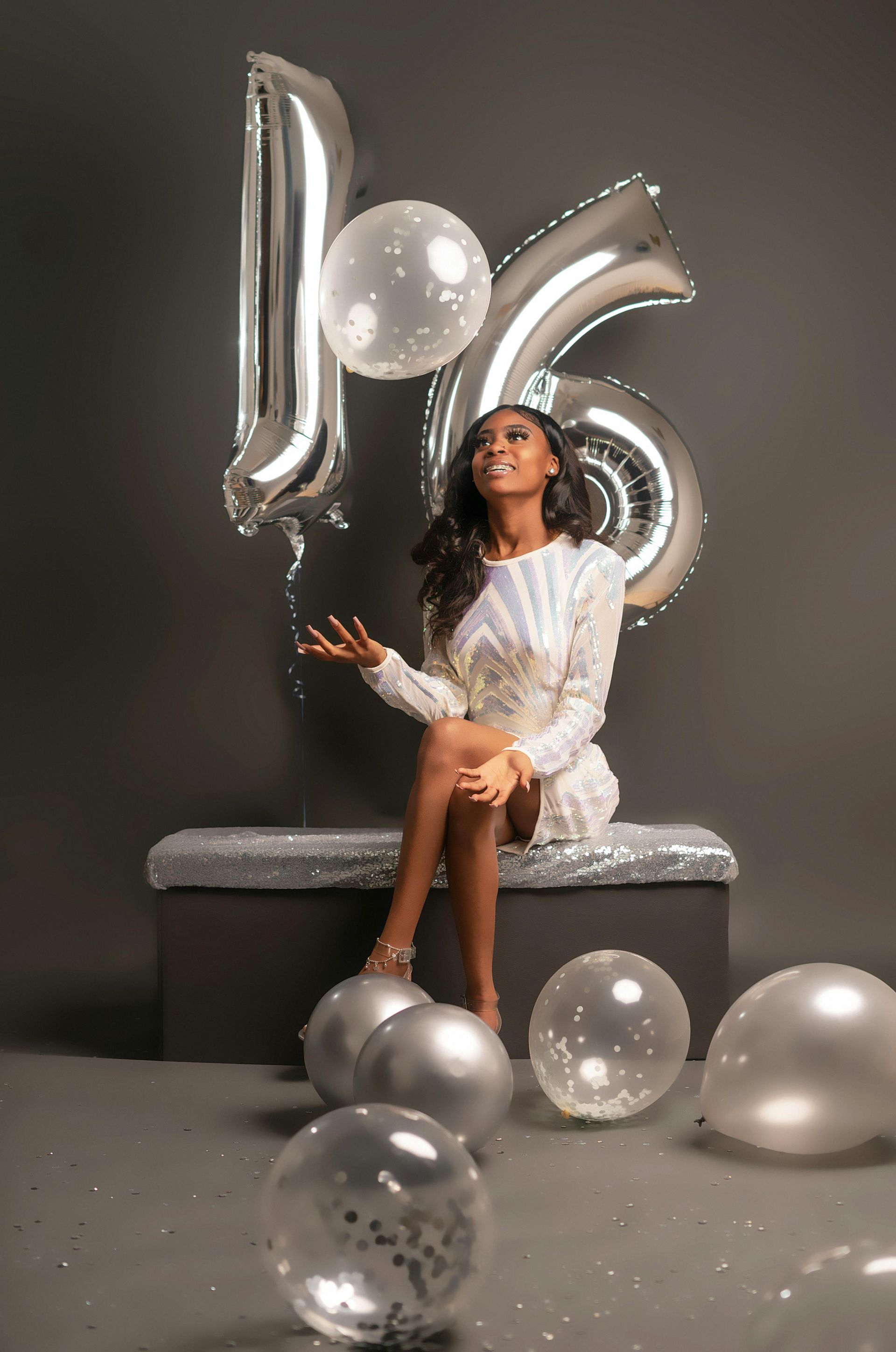 Woman in white dress seated on a silver bench with silver balloons and metallic number balloons against a dark backdrop