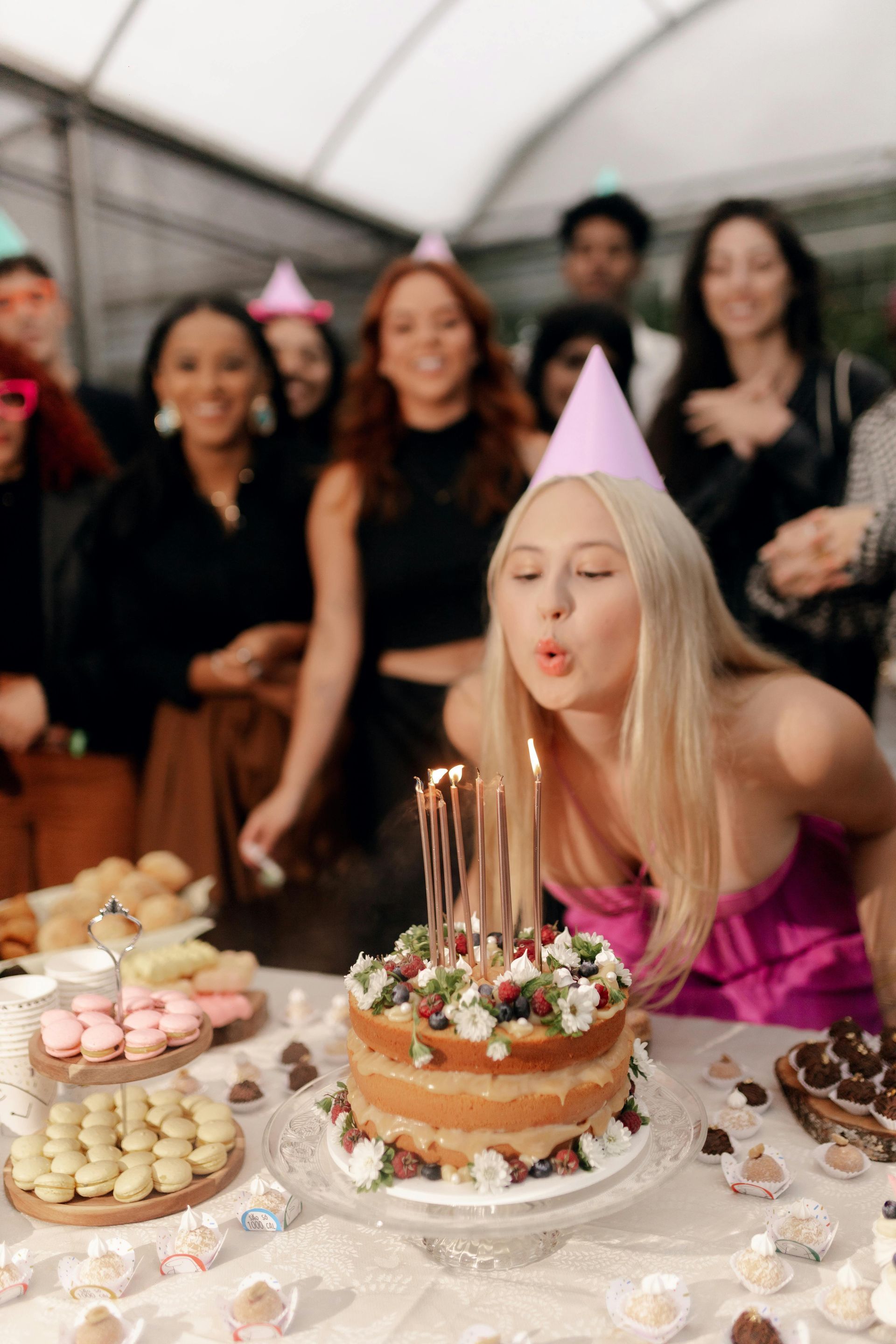 Woman in a pink party hat blowing out candles on a decorated birthday cake at a celebration