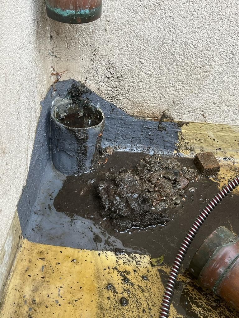 Coin de sol sale avec un drain endommagé, de l’eau stagnante et des débris près de tuyaux et d’un mur.