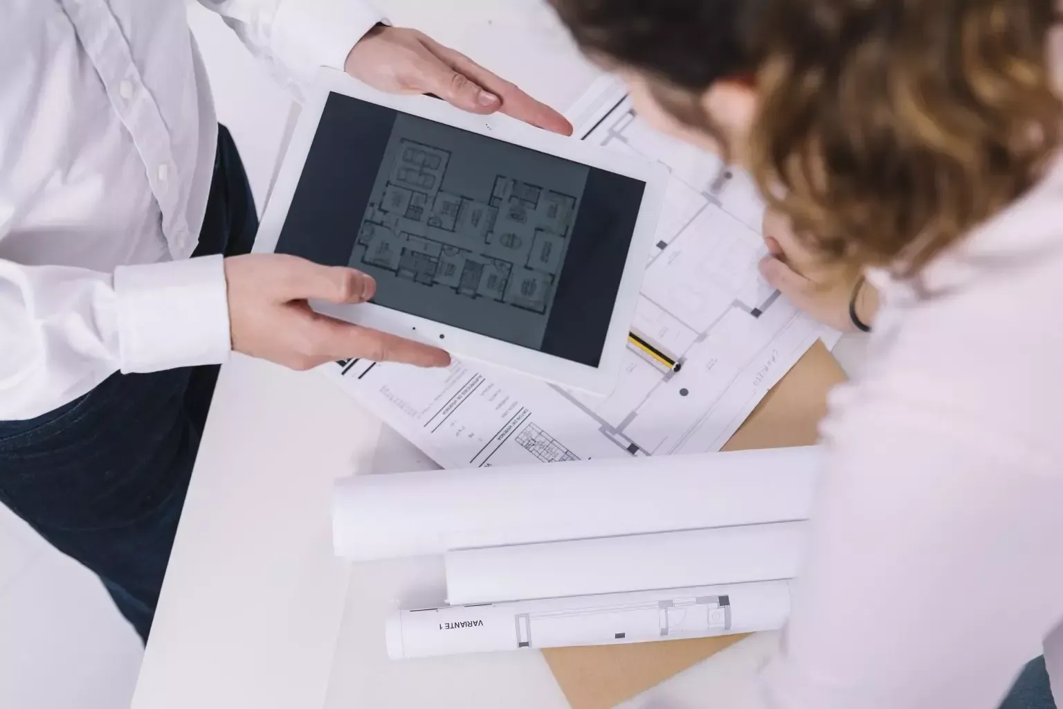 Duas pessoas examinam um tablet sobre desenhos arquitetônicos em uma mesa.