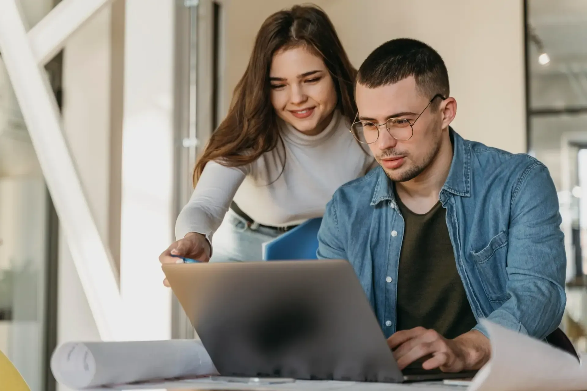 Duas pessoas colaborando em um laptop em um escritório iluminado, revisando algo juntas e sorrindo.