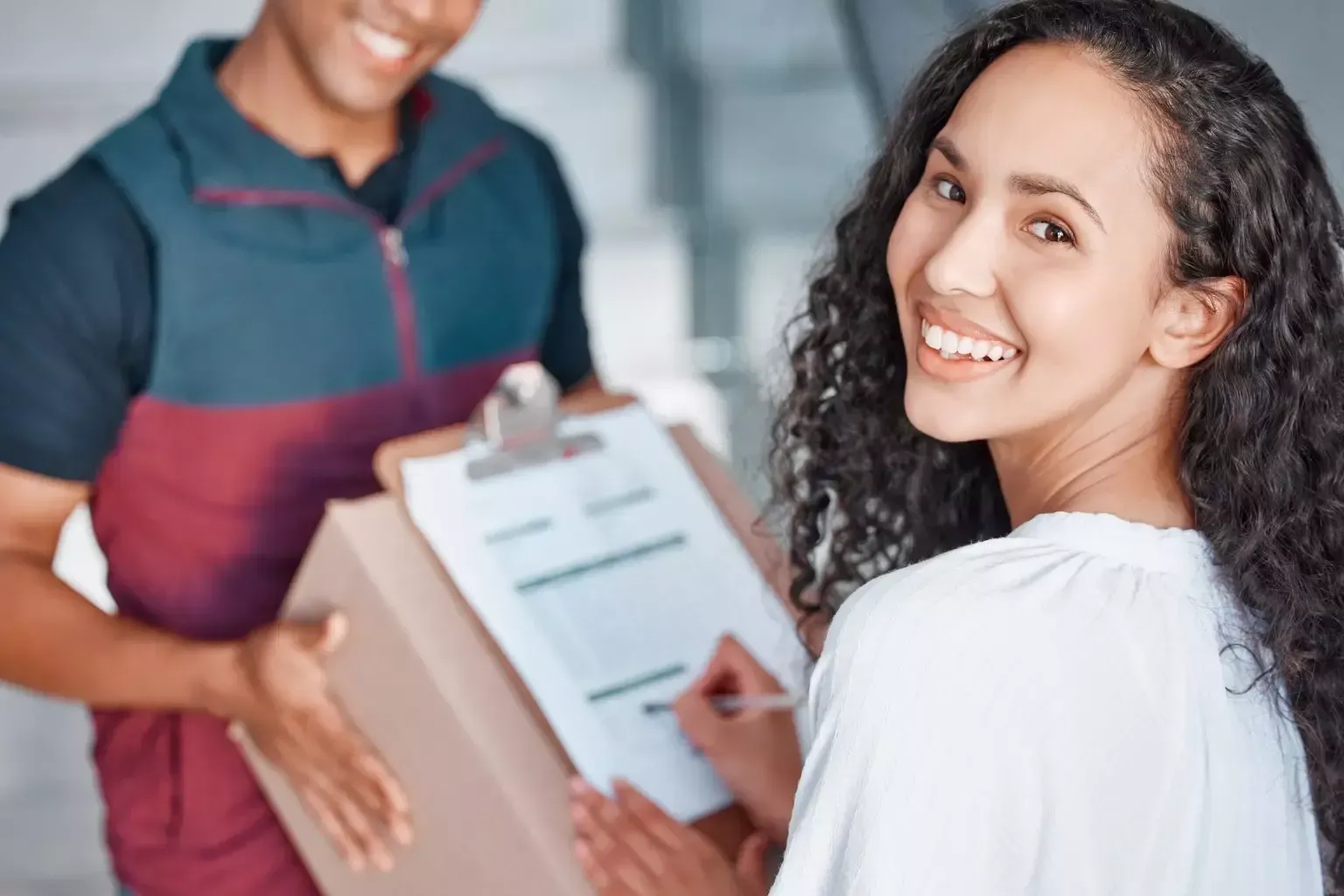 Pessoa sorridente segurando uma prancheta enquanto entrega um formulário para outra pessoa em ambiente interno.