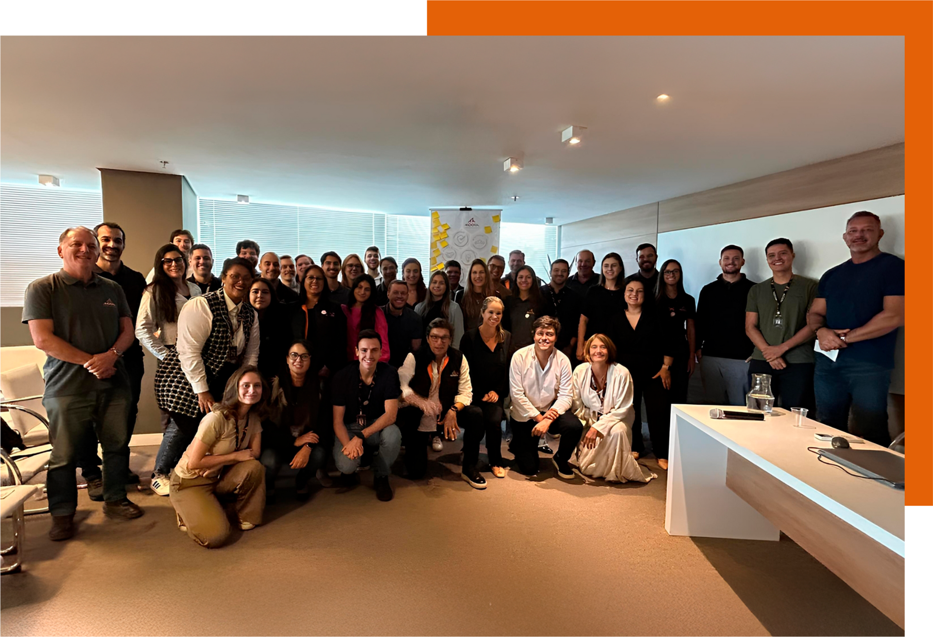 Um grande grupo posando em uma sala de reuniões interna, com iluminação aconchegante e uma mesa branca à direita.
