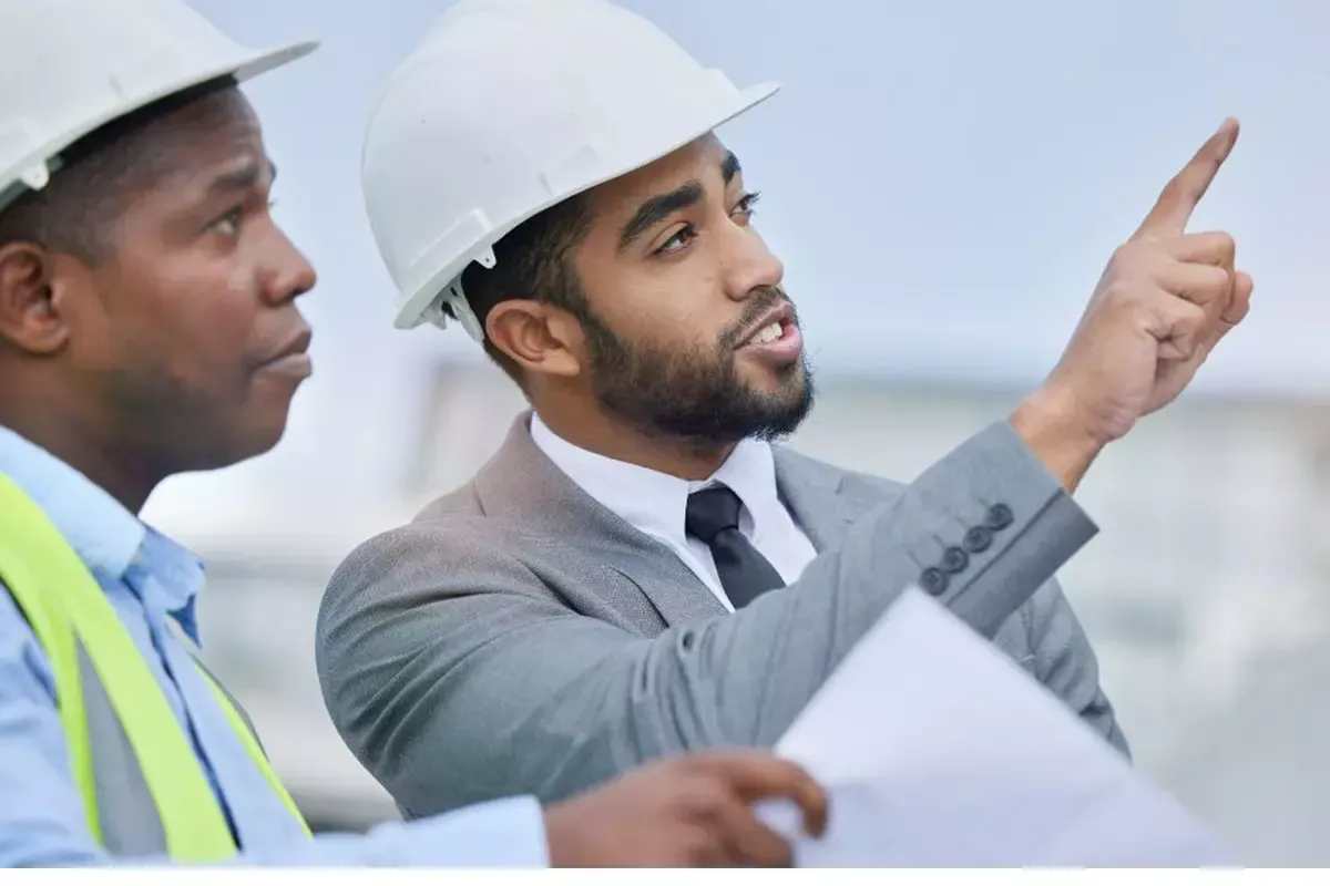 Dois operários da construção civil com capacetes revisando plantas, um deles apontando para a frente.