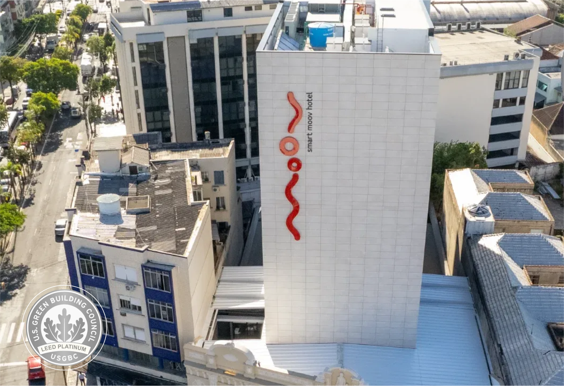 Vista aérea de um edifício hospitalar branco com um logotipo vertical vermelho em uma área urbana.